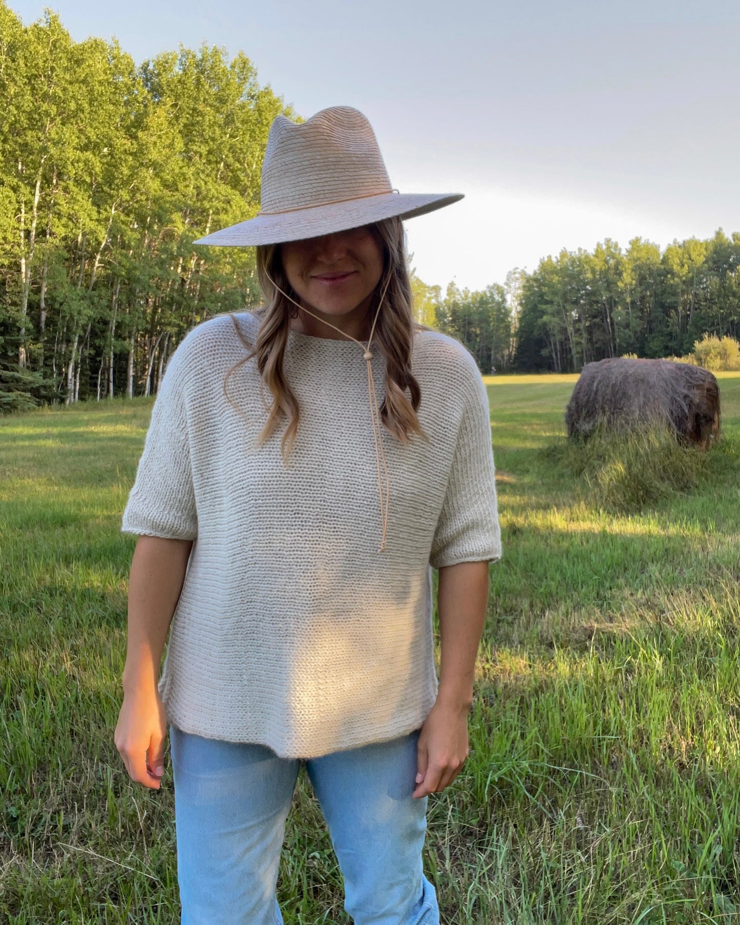 Person wearing a wide-brimmed hat and light sweater standing in a field with trees and hay bales in the background.