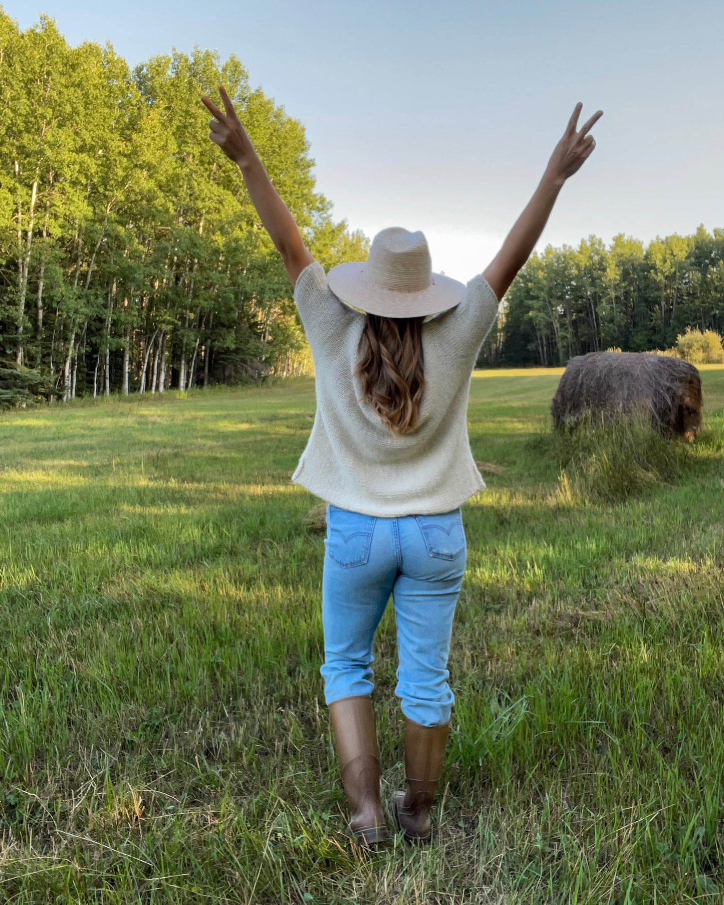 Person standing in a field with arms raised, wearing a hat and knit t-shirt.