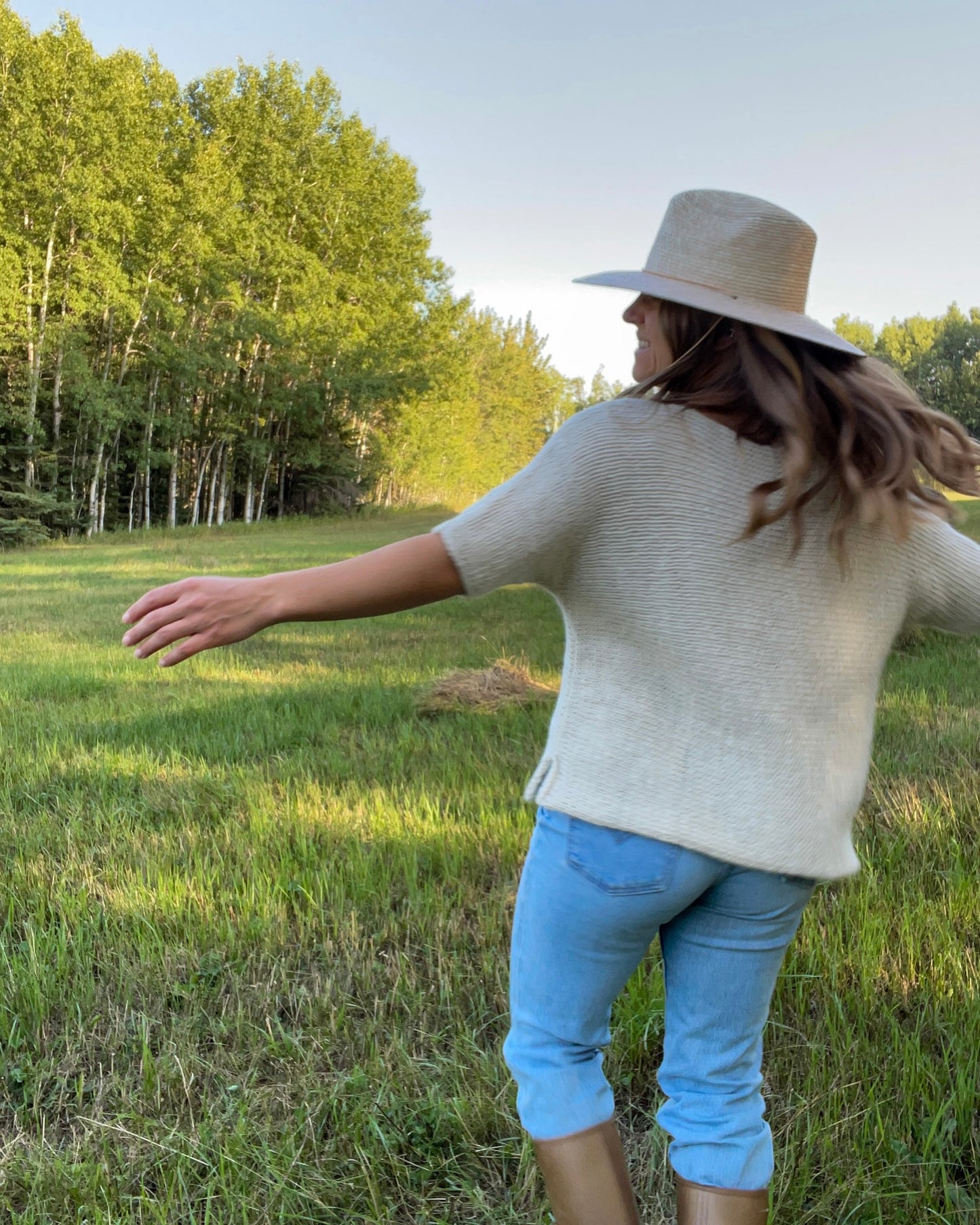 Person wearing a white knit t-shirt and blue jeans standing in a grassy field with trees in the background