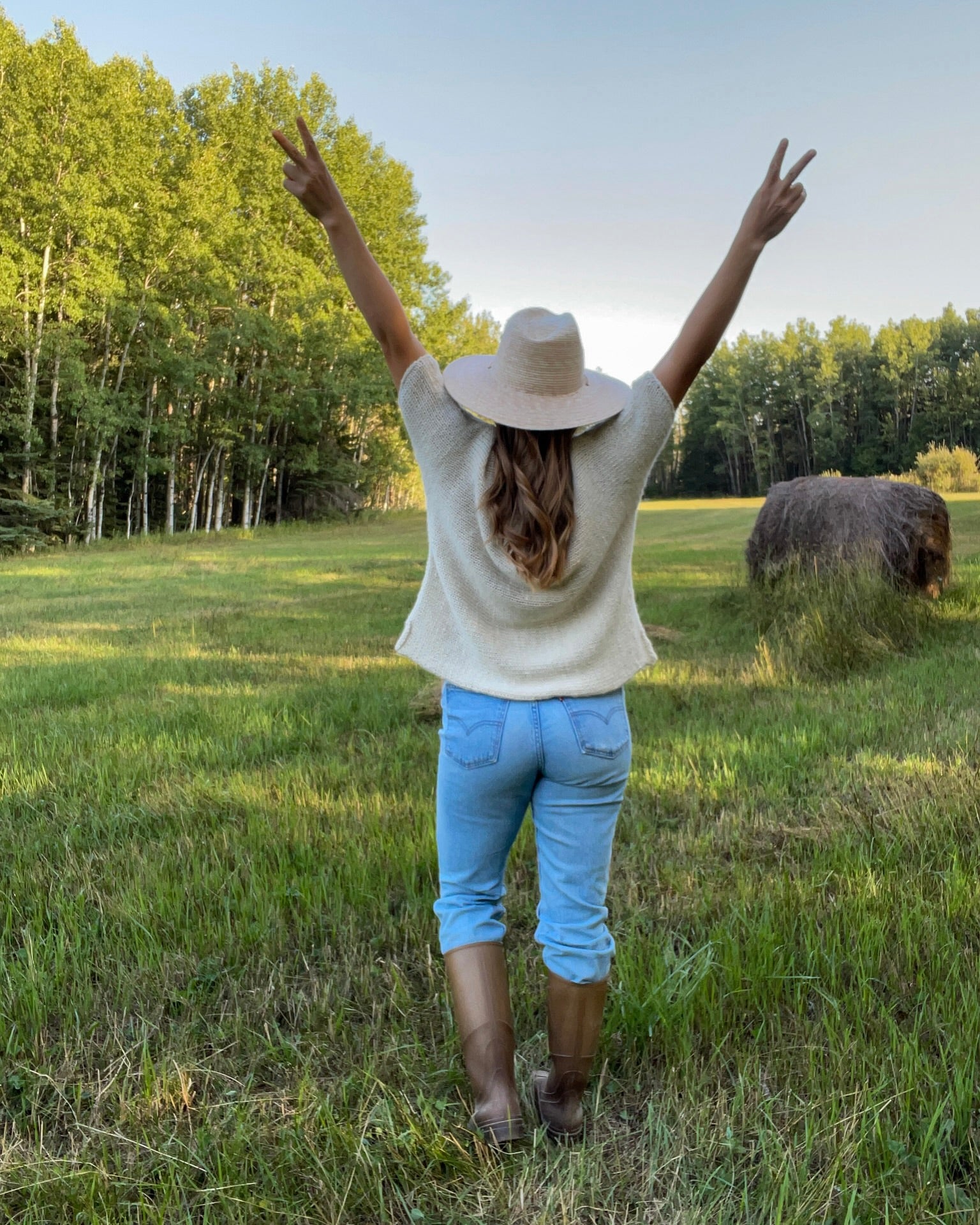 Person standing in a field with arms raised, wearing a hat and knit t-shirt.