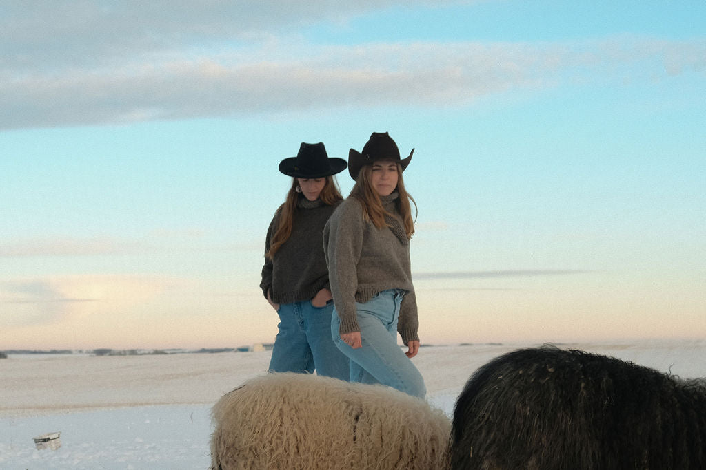 Two women in cowboy hats standing next to a sheep on a snowy landscape.