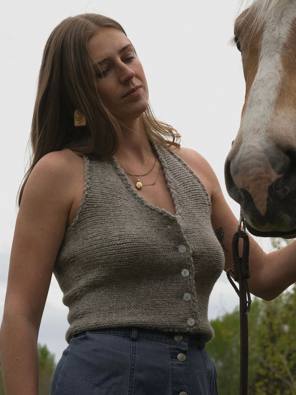 Woman wearing a sleeveless knitted top standing next to a horse outdoors.
