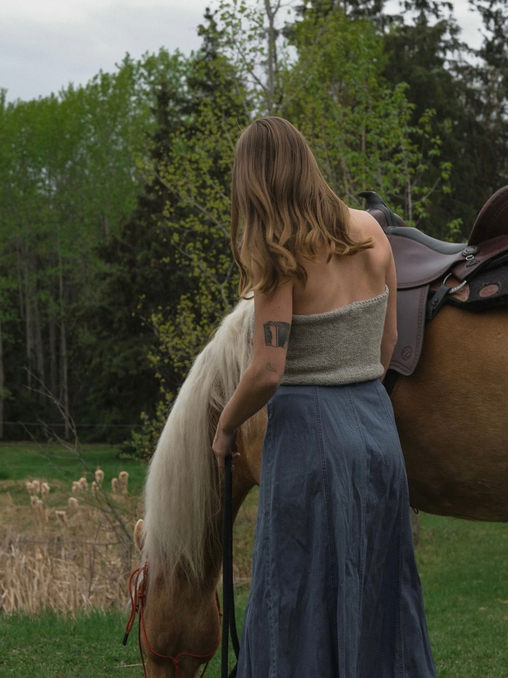 Woman wearing a sleeveless knitted top standing next to a horse outdoors.