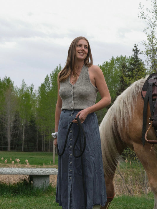 Woman wearing a sleeveless knitted top standing next to a horse outdoors.
