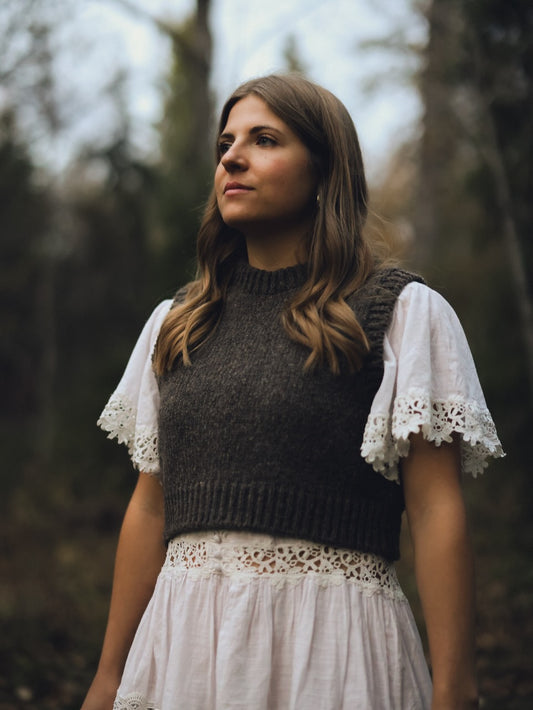 Woman wearing a dark knit top and white lace-trimmed dress in a forest setting