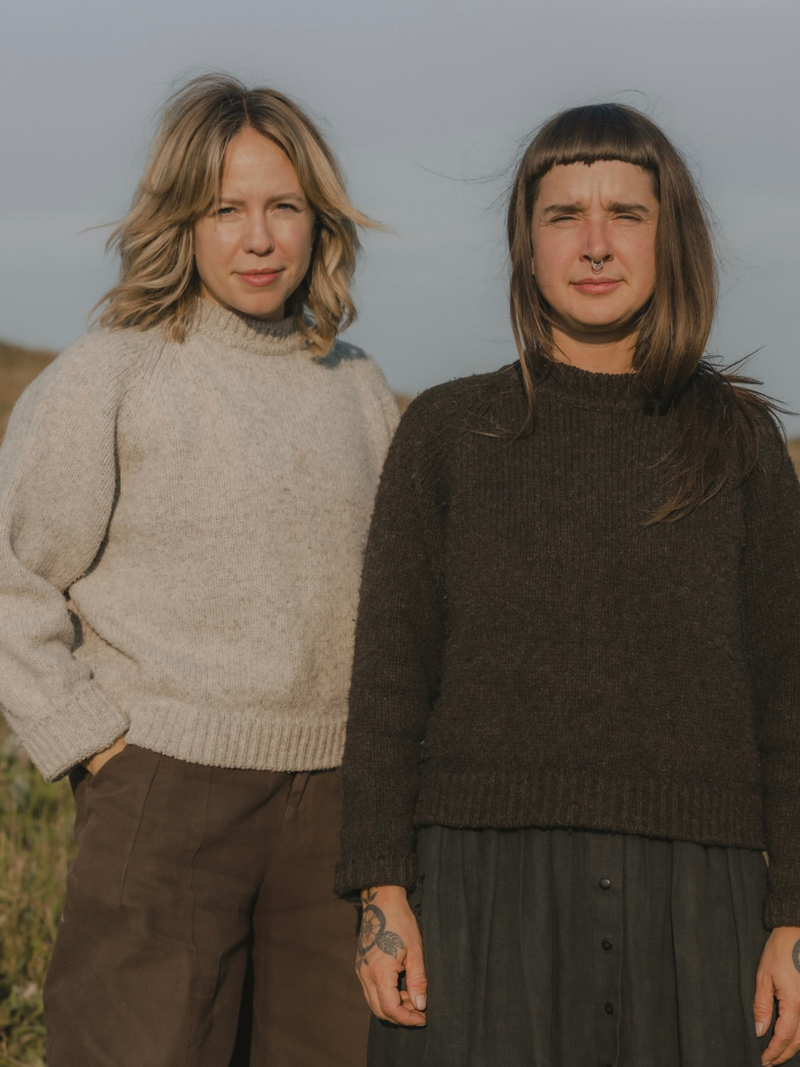 Two women standing outdoors in a field, wearing cozy knit sweaters. The woman on the left wears a light grey sweater and brown pants.