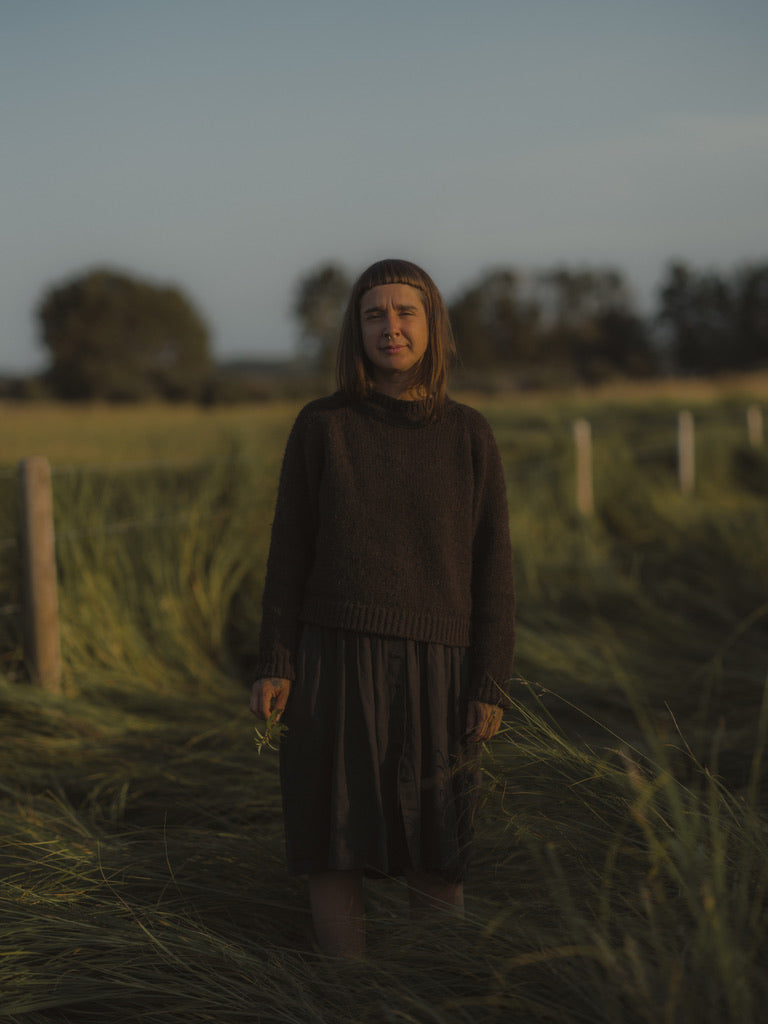 A woman stands in a grassy field at dusk wearing a dark brown wool sweater and a dark skirt.