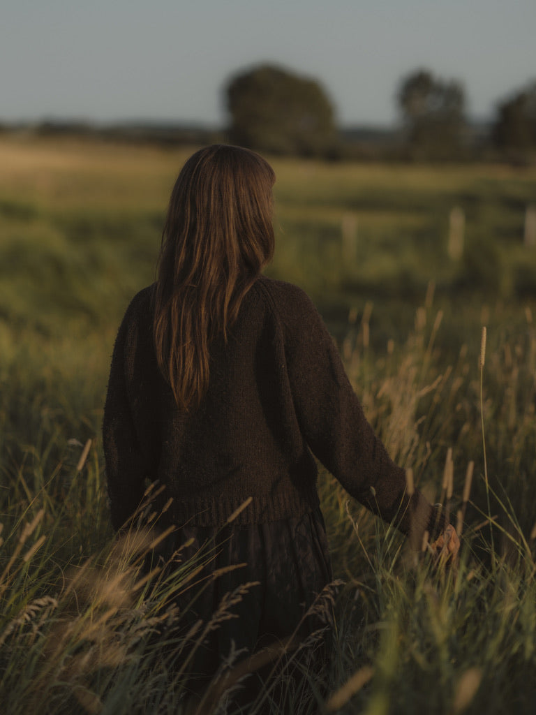 A woman with long light brown hair seen from behind, wearing a dark brown knit sweater and dark skirt, standing in a grassy field at sunset.
